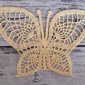 Puede incluir: Una mariposa de ganchillo de color amarillo p&aacute;lido. La mariposa tiene detalles intrincados y un dise&ntilde;o delicado.