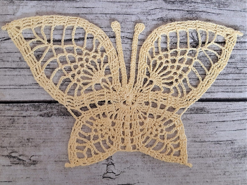 Puede incluir: Una mariposa de ganchillo de color amarillo p&aacute;lido. La mariposa tiene detalles intrincados y un dise&ntilde;o delicado.