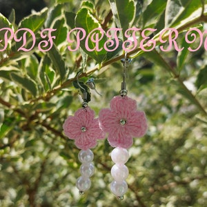 May include: Pink crocheted flower earrings with white pearl beads hanging from the bottom. The earrings are hanging from a branch with green leaves.