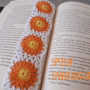 May include: Crocheted bookmark featuring a repeating pattern of orange and yellow flower designs with white borders. The bookmark is placed on an open book, enhancing its visual appeal.