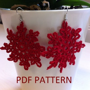 Pode incluir: Um par de brincos de floco de neve de crochê vermelho pendurados em ganchos prateados. Os brincos são exibidos em um vaso de cerâmica branco com o texto "PDF PATTERN" impresso abaixo.