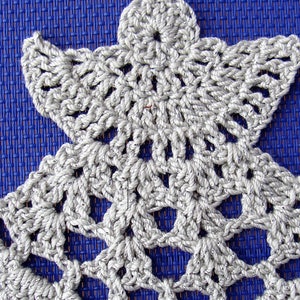 Puede incluir: Un adorno de &aacute;ngel de crochet blanco con un dise&ntilde;o detallado. El adorno est&aacute; sobre un fondo azul.