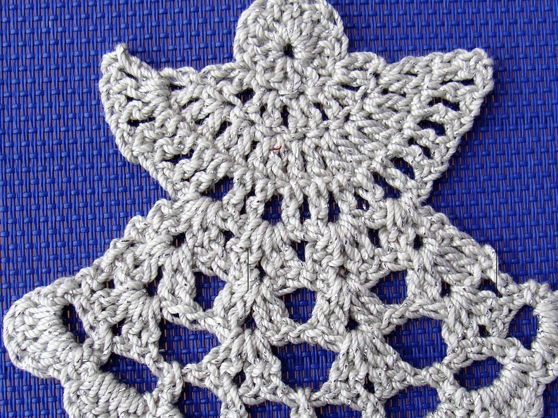 Puede incluir: Un adorno de &aacute;ngel de crochet blanco con un dise&ntilde;o detallado. El adorno est&aacute; sobre un fondo azul.