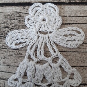Puede incluir: Adorno de &aacute;ngel blanco tejido a crochet con un dise&ntilde;o de flor en la parte superior y una falda en forma de campana. El adorno est&aacute; sobre un fondo marr&oacute;n texturizado.