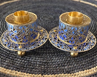 Könnte beinhalten: Zwei verzierte silberne Kerzenhalter mit blauen und goldenen Emaille-Designs. Jeder Halter hat eine runde Basis und eine zentrale Tasse für eine Kerze. Die Halter stehen auf einer schwarzen geflochtenen Oberfläche.
