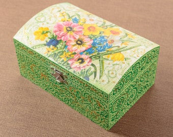 Puede incluir: Una caja de madera verde con un diseño floral en la tapa. La caja tiene una tapa con bisagras y un cierre de metal. El diseño floral presenta flores rosas, amarillas y blancas.