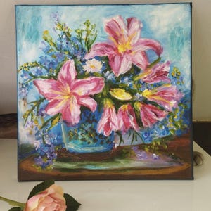 May include: Oil painting of a floral arrangement featuring pink lilies and blue wildflowers in a blue vase. The artwork is on a square canvas with a light blue background. A pink rose lies in the foreground.