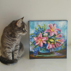 May include: An oil painting of pink lilies and blue flowers in a blue vase. The artwork is on a square canvas. A grey tabby cat sits beside the painting.