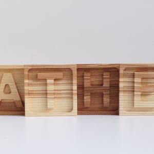May include: Wooden blocks spelling out the word "FATHER" in all capital letters. Each block is a different shade of brown wood.