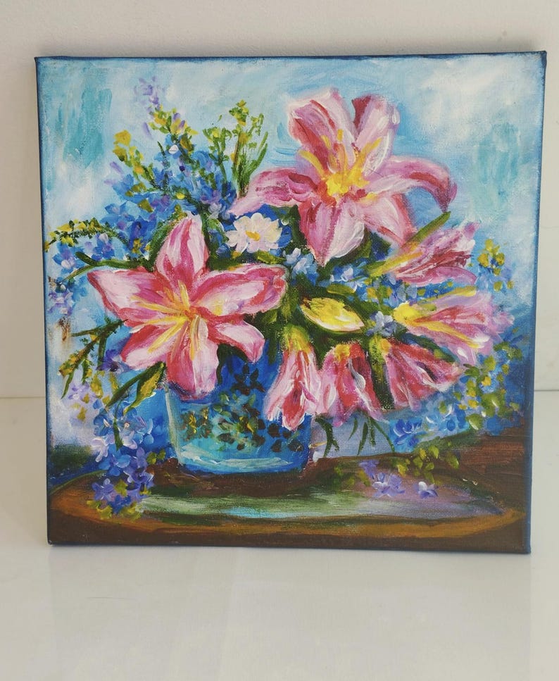 May include: Oil painting depicting a floral arrangement. Pink lilies with yellow centres and blue forget-me-nots are arranged in a blue vase. The painting is set against a light blue background, with a wooden surface.