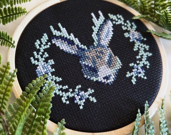 May include: A black embroidery hoop with a blue and white cross-stitch design of a rabbit with antlers surrounded by a wreath of leaves. The hoop is on a white surface.