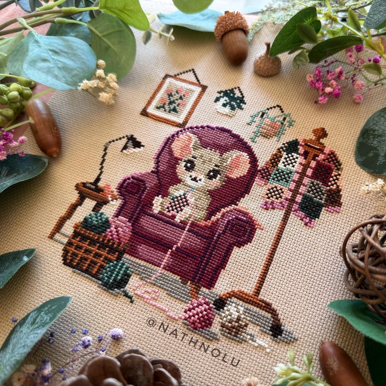Modern cross stitch design of a cute mouse crocheting granny squares in a cozy cottagecore setting