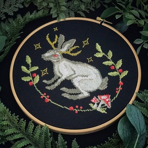 May include: A white rabbit with antlers and a red eye is embroidered on a black fabric with a wooden hoop. The rabbit is surrounded by green leaves and red berries.