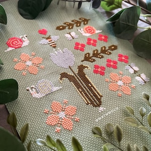 May include: A cross-stitch pattern featuring a green background with a floral design. The design includes pink flowers, green leaves, white tulips, and brown stems. There are also snails, butterflies, and bees in the design.
