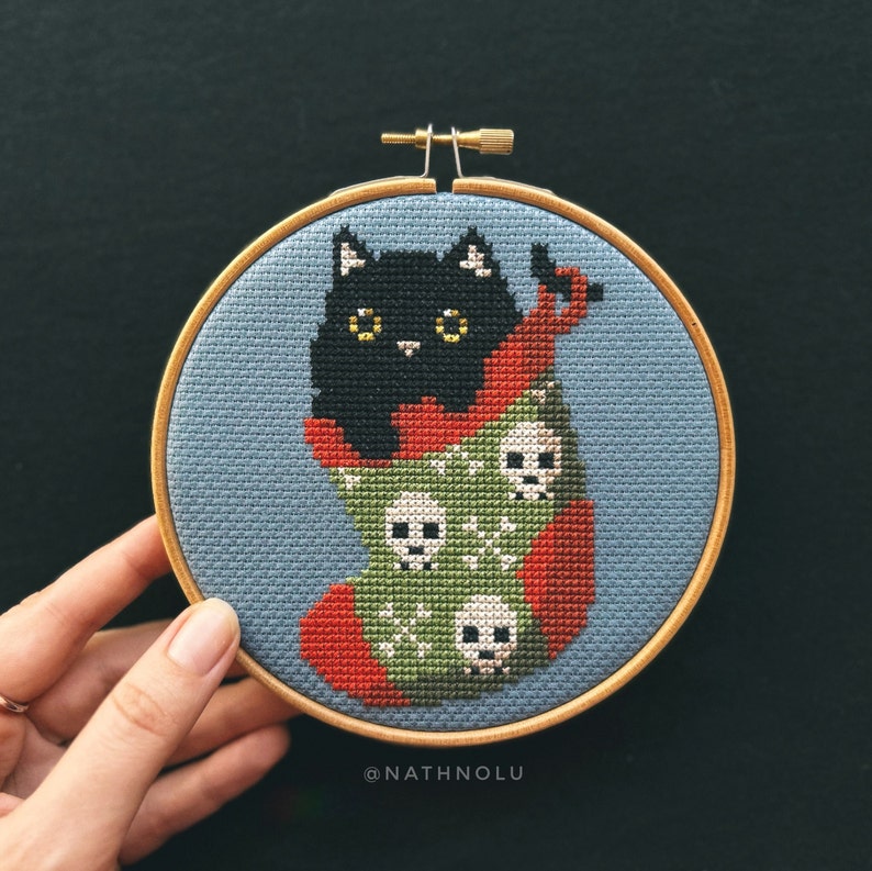 May include: A black cat with yellow eyes sits inside a red and green stocking with a skull pattern. The stocking is in a wooden embroidery hoop.