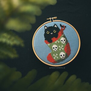 May include: A black cat cross-stitched in a red and green stocking with white skulls. The stocking is in a blue embroidery hoop with the text "@NATHNOLU" below.