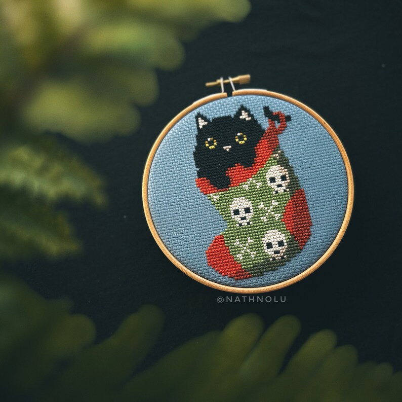 May include: A black cat cross-stitched in a red and green stocking with white skulls. The stocking is in a blue embroidery hoop with the text "@NATHNOLU" below.