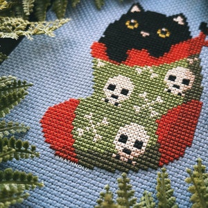 May include: A black cat peeking out of a green and red stocking with white skulls. The stocking is on a blue background.