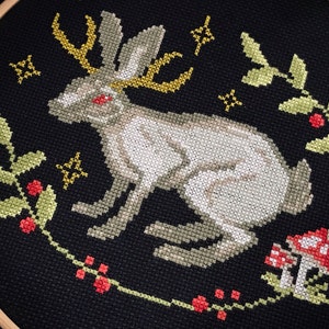 Cross stitch horned hare on black aida surrounded by plants with red berries and fly agarics. over the horns of the hare small stars