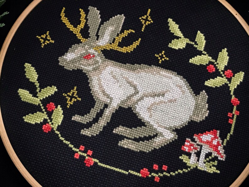 Cross stitch horned hare on black aida surrounded by plants with red berries and fly agarics. over the horns of the hare small stars
