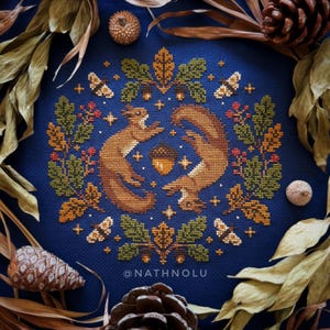 May include: A cross-stitch embroidery on dark blue fabric featuring two brown squirrels facing each other, surrounded by a circular wreath of green and gold oak leaves, red berries, and small moths. A single acorn sits in the centre. Text reads @NATHNOLU.
