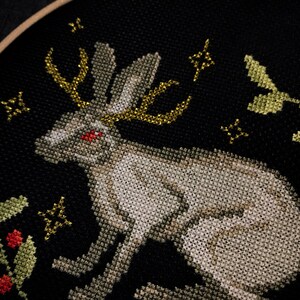 Jacalope cross stitch on black aida. The photo shows how the horns shimmer when the light hits them. Around the hare is a plant ornament