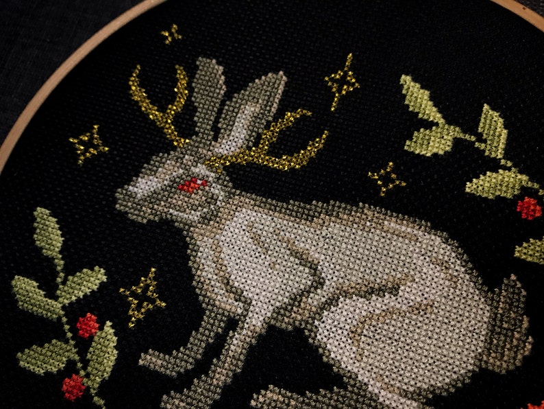 Jacalope cross stitch on black aida. The photo shows how the horns shimmer when the light hits them. Around the hare is a plant ornament