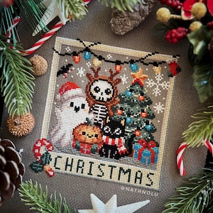 以下が含まれることがあります： 骸骨のトナカイ、幽霊、カボチャ、黒猫、プレゼントをつけたクリスマスツリーが描かれたクロスステッチのパターン。デザインは額装されており、「CHRISTMAS」と「@NATHNOLU」の文字が含まれています。