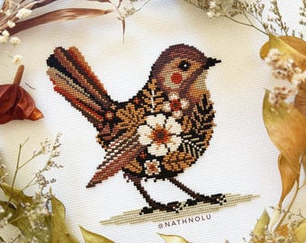 May include: A detailed cross-stitch of a bird with brown, black, and orange hues, featuring floral patterns on its body. The bird is set against a white background, surrounded by dried leaves and flowers. The artist's name, @NATHNOLU, is visible.