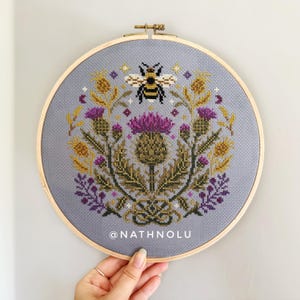 Nature-inspired cross stitch pattern with blooming thistles, wildflowers, and a detailed bumblebee embroidery design.