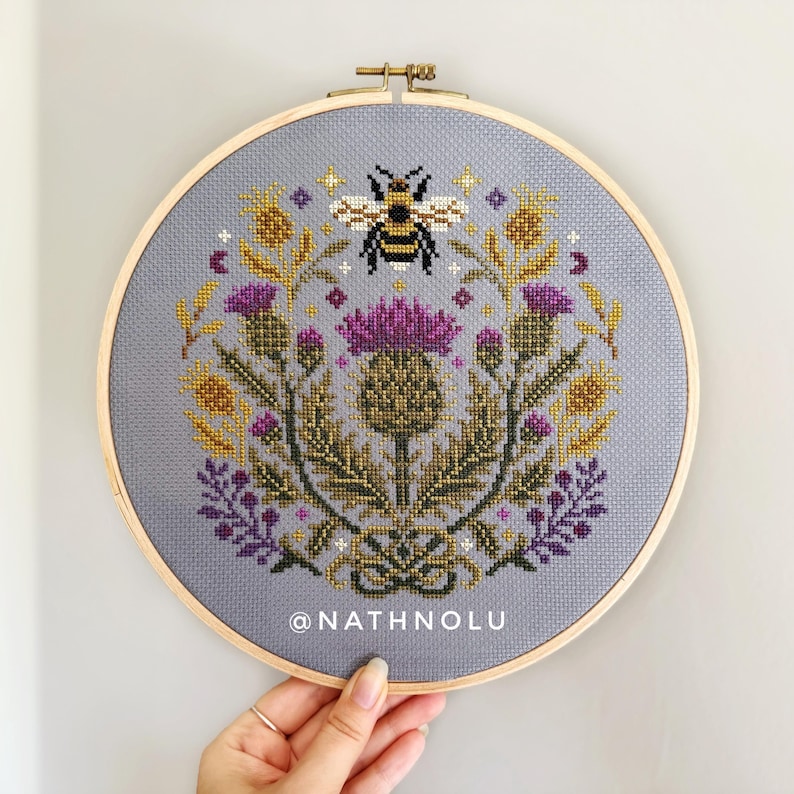 Nature-inspired cross stitch pattern with blooming thistles, wildflowers, and a detailed bumblebee embroidery design.