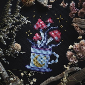 以下が含まれることがあります： 三日月と星が描かれた青いマグカップに、ピンクと赤のキノコが詰まっているクロスステッチ刺繍のデザイン。デザインは黒い背景に囲まれており、下部に「@NATHNOLU」というテキストがあります。