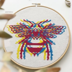 May include: A colorful cross-stitch embroidery of a bee with yellow, red, and blue accents. The bee is framed in a wooden hoop.