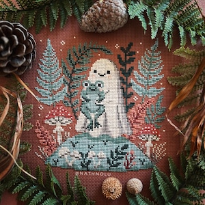 Könnte beinhalten: Kreuzstichmuster mit einem weißen Geist, der einen grünen Frosch hält, umgeben von Farnen und Pilzen auf braunem Hintergrund. Das Muster enthält den Text "@NATHNOLU".