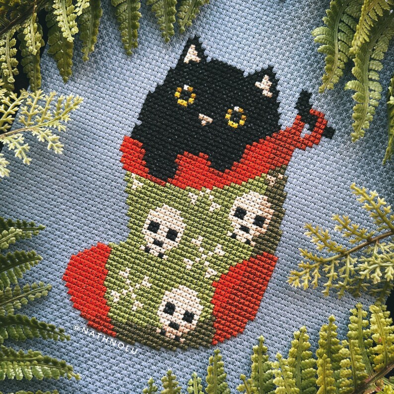 May include: A black cat with yellow eyes sits inside a green and red stocking with skulls embroidered on it. The stocking is filled with the cat and has a red cuff.