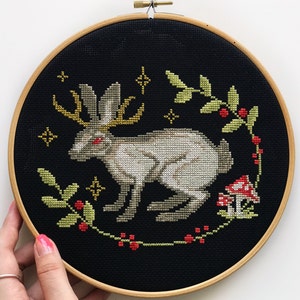 Cross embroidery of a mythical Easter bunny with horns on black canvas in an embroidery frame, holding his hand against a light background