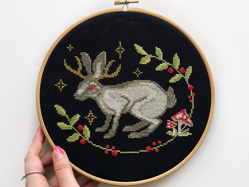 Cross embroidery of a mythical Easter bunny with horns on black canvas in an embroidery frame, holding his hand against a light background