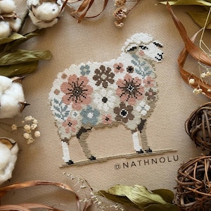 Peut inclure: Une broderie au point de croix sur tissu beige représentant un mouton orné d'un motif floral dans des tons rose, marron et bleu. Le mouton a une tête et des pattes blanches, et le texte @NATHNOLU est brodé dans le coin inférieur droit.