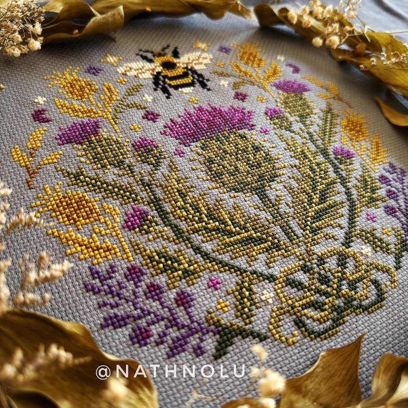 Close-up of the bee embroidery showing detailed wings and floral thistle motifs in a repeating cross stitch pattern.