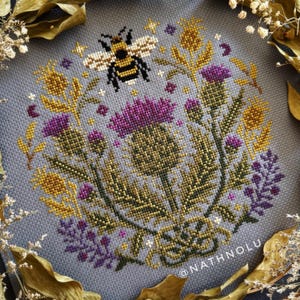 Botanical cross stitch design with purple thistle flowers and a bee, inspired by wildflower meadows and cottagecore decor.