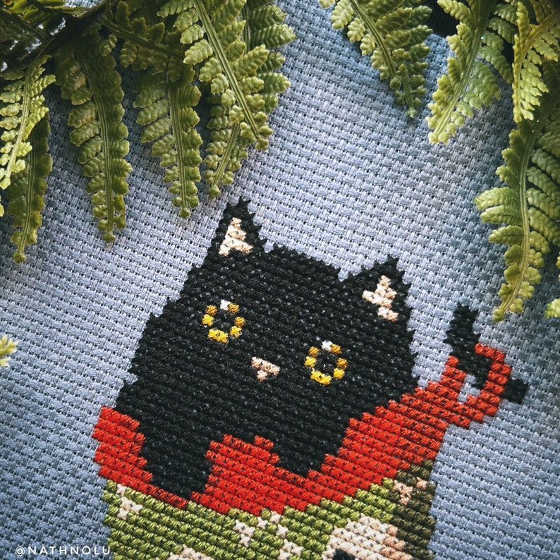 May include: Cross-stitch pattern of a black cat wearing a red and green sweater. The cat is looking directly at the viewer with yellow eyes.