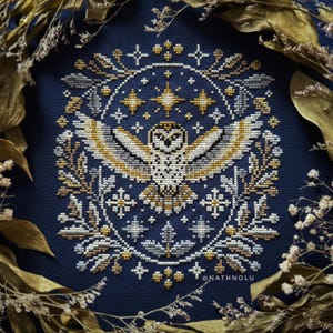 Peut inclure: Une broderie au point de croix représentant un hibou aux ailes déployées, entouré d'une couronne de feuilles, d'étoiles et d'éléments floraux. Le motif utilise du fil doré, blanc et gris sur un fond bleu marine. Le texte "@nathnolu" est visible.