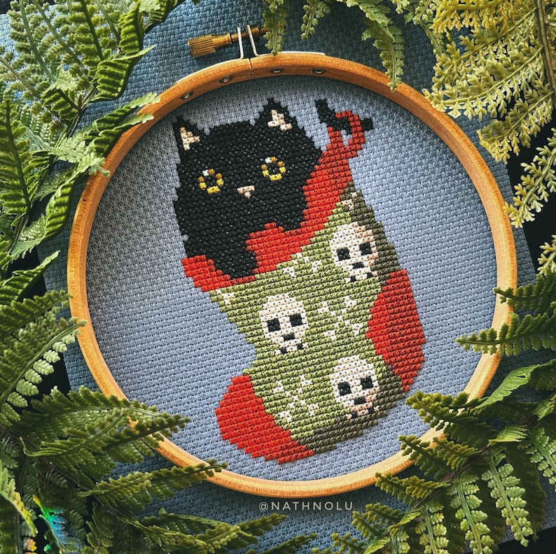 May include: A black cat peeking out of a green and red stocking with white skull designs. The stocking is framed in a wooden hoop with a blue background. The text "@NATHNOLU" is at the bottom of the hoop.
