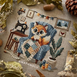 Cozy woodland-themed cross stitch pattern featuring a fox crafting a quilt in a blue chair.