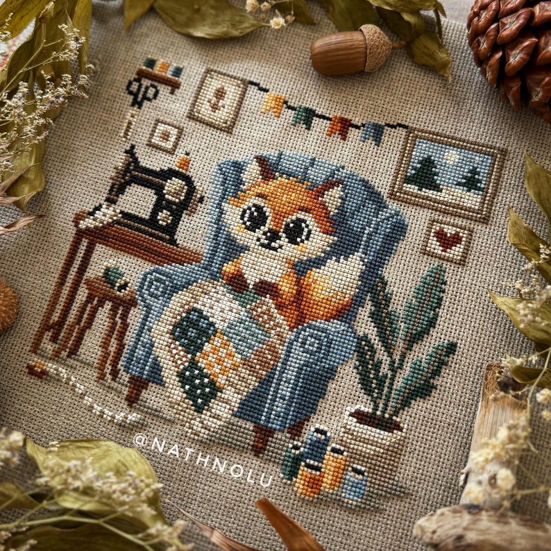 Cozy woodland-themed cross stitch pattern featuring a fox crafting a quilt in a blue chair.