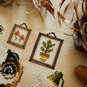 Wall decor details from the Reading Badger cross stitch design showing framed pictures of plants and mushrooms.