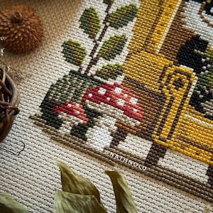 Cozy farmhouse cross stitch of a reading badger and frog with books, plants and mushrooms, warm autumn tones.