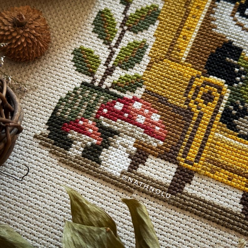 Cozy farmhouse cross stitch of a reading badger and frog with books, plants and mushrooms, warm autumn tones.