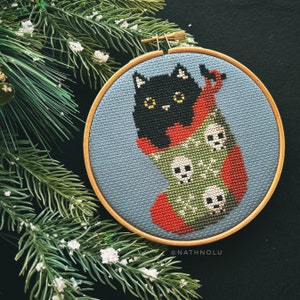 May include: A black cat with yellow eyes peeks out of a green and red stocking with white skull designs. The stocking is embroidered on a blue fabric background and framed in a wooden hoop. The text "@NATHNOLU" is at the bottom of the hoop.