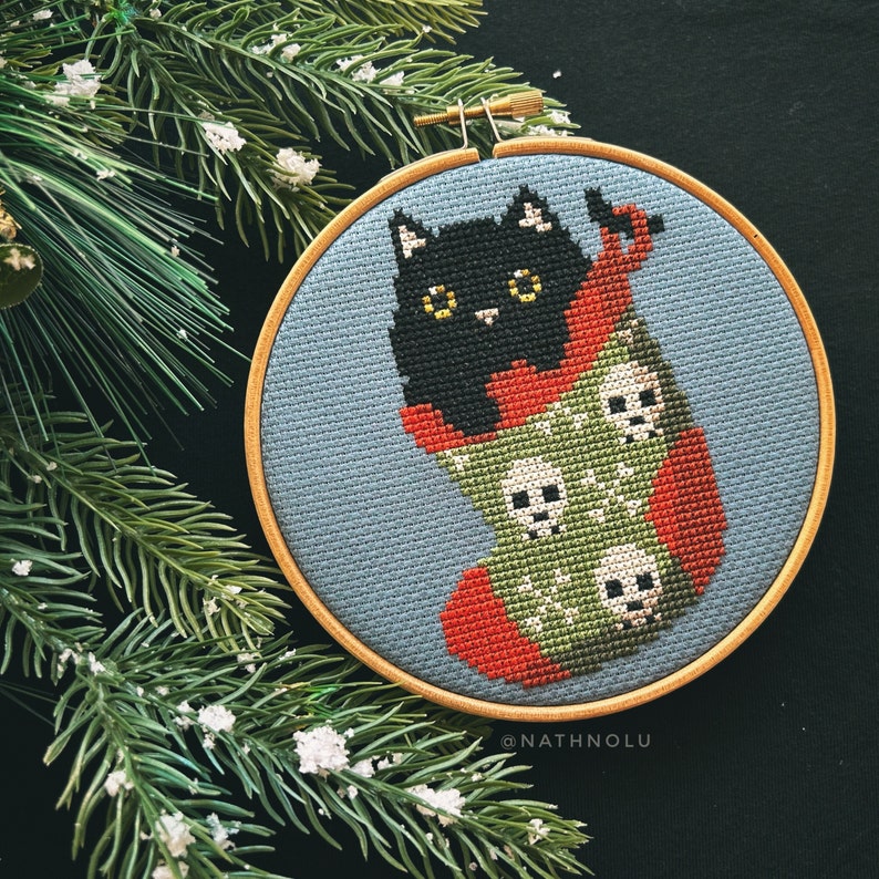 May include: A black cat with yellow eyes peeks out of a green and red stocking with white skull designs. The stocking is embroidered on a blue fabric background and framed in a wooden hoop. The text "@NATHNOLU" is at the bottom of the hoop.